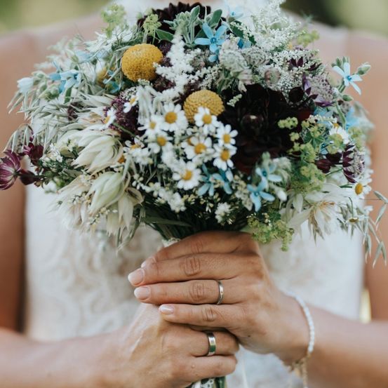 Eine Braut hält einen Hochzeitsstrauß mit weißen, blauen und gelben Blumen und grünen Blättern.
