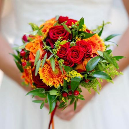Eine Braut in einem weißen Hochzeitskleid hält einen Brautstrauß aus roten Rosen, orangefarbenen und gelben Gänseblümchen und grünen Blättern.