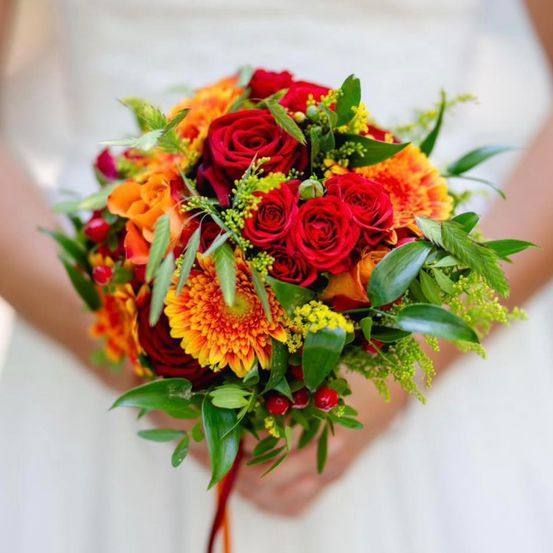 Eine Braut in einem weißen Hochzeitskleid hält einen Brautstrauß aus roten Rosen, orangefarbenen und gelben Gänseblümchen und grünen Blättern.