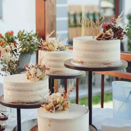 Vier Hochzeitstorten mit weißer Glasur und verschiedenen Blumendekorationen stehen auf Holzständern in einer Gartenumgebung.