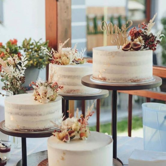 Vier Hochzeitstorten mit weißer Glasur und verschiedenen Blumendekorationen stehen auf Holzständern in einer Gartenumgebung.