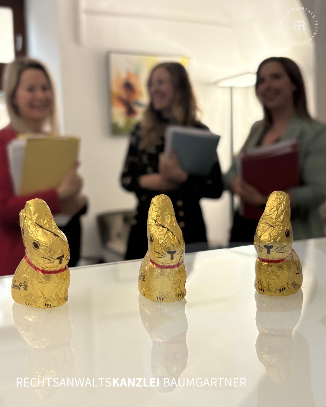 Drei goldene Schokoladen-Osterhasen stehen auf einem Tisch. Hinter den Hasen lächeln drei Frauen und halten Ordner. Die Frau links trägt ein rotes Oberteil, die Frau in der Mitte ein schwarzes Oberteil und die Frau rechts einen grünen Blazer.