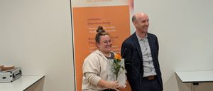 Eine Frau und ein Mann stehen lächelnd vor einem Connexia-Banner. Die Frau hält einen Blumenstrauß und ein Zertifikat.