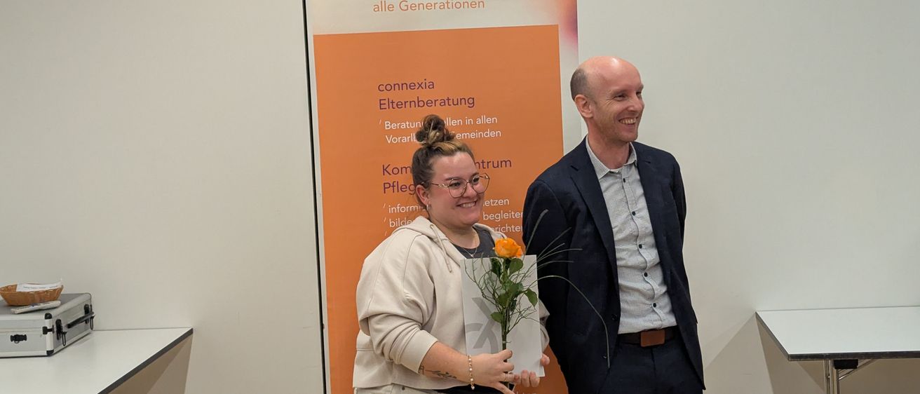 Eine Frau und ein Mann stehen lächelnd vor einem Connexia-Banner. Die Frau hält einen Blumenstrauß und ein Zertifikat.