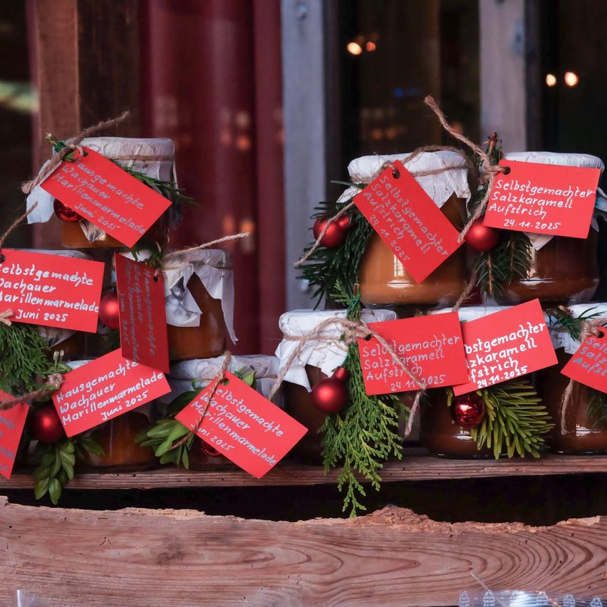 Ein Holztisch trägt Gläser mit hausgemachter Marmelade, jedes mit einem roten Etikett, das den Hersteller und das Datum angibt. Grüne Girlanden mit roten Ornamenten schmücken die Gläser, vor einem unscharfen Hintergrund.