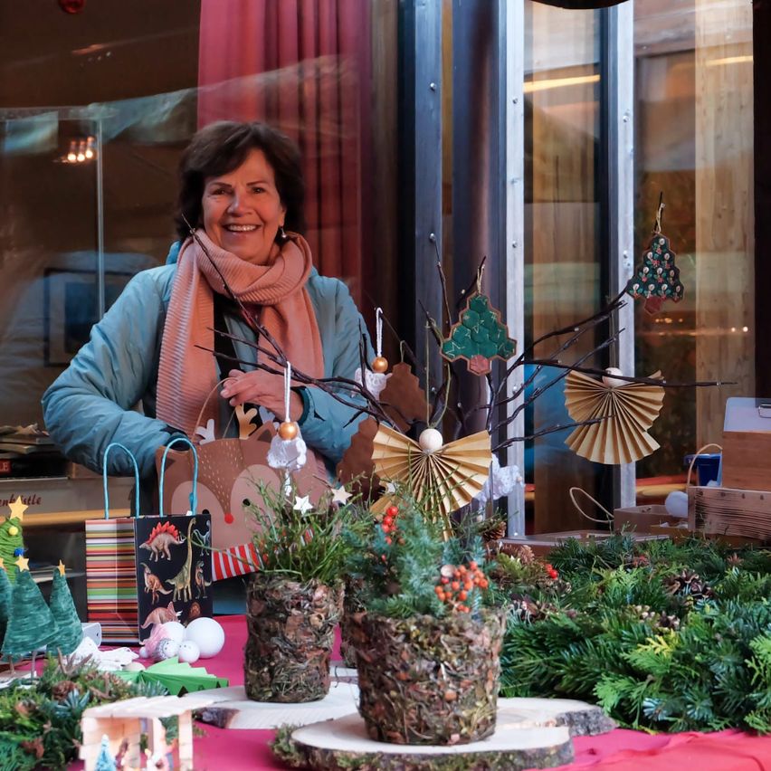 Eine Frau mit einem Schal steht hinter einem Tisch mit Weihnachtsdekoration. Der Tisch hat zwei Vasen mit Pflanzen, Geschenktüten und anderen festlichen Gegenständen. Hinter ihr befindet sich ein Fenster mit einer Reflexion eines Gebäudes.
