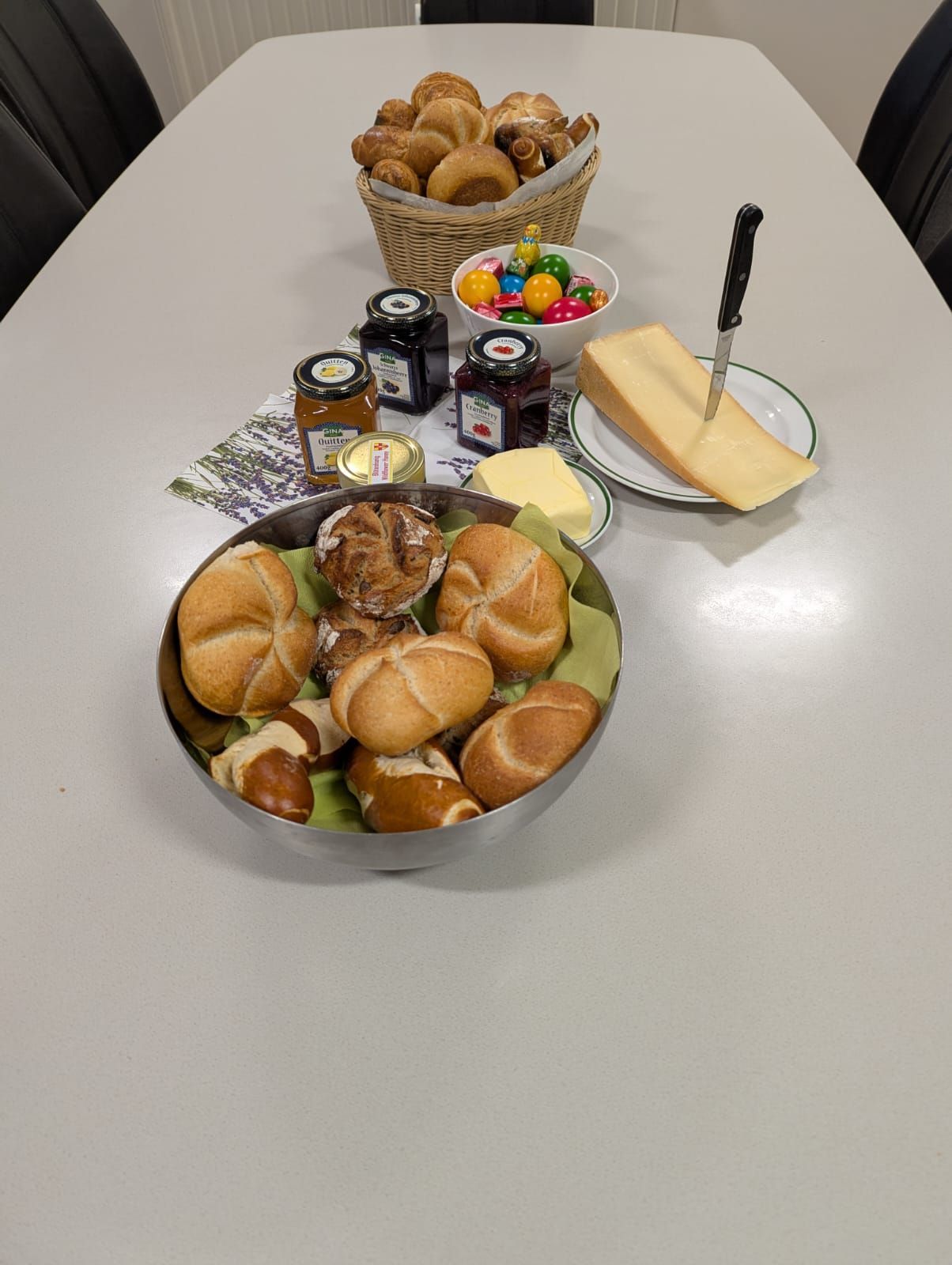 A white table with a bowl of bread rolls, a basket of bread, cheese, butter, jam, and Easter eggs. A knife and a plate with cheese are also on the table.