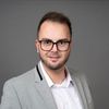Ein Mann in einem grauen Blazer und einem weißen Hemd mit schwarzer Brille posiert für ein Foto vor einem grauen Hintergrund.