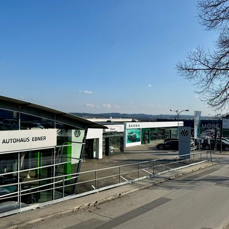 Außenansicht eines Autohauses unter einem klaren blauen Himmel. Das große Schild lautet Autohaus Ebner. Ein Skoda-Auto ist draußen ausgestellt.