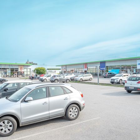 Ein Parkplatz im Freien mit mehreren geparkten Autos. Einige Autos sind in der Nähe der Geschäfte geparkt, während andere auf dem Parkplatz geparkt sind.