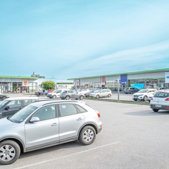 Ein Parkplatz im Freien mit mehreren geparkten Autos. Einige Autos sind in der Nähe der Geschäfte geparkt, während andere auf dem Parkplatz geparkt sind.