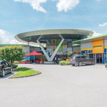 Außenansicht eines Einkaufszentrums. Eine Person geht auf dem Gehweg, und ein roter Schirm steht draußen. Autos sind auf dem Parkplatz geparkt.