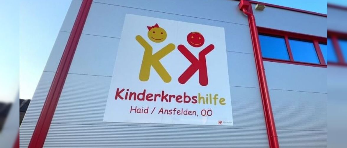 Ein Gebäudeaußenbereich mit einem Schild, das 'KK Kinderkrebshilfe Haid / Ansfelden, OO' besagt. Das Schild zeigt zwei lächelnde Gesichter und ist über einem roten Rohr angebracht.
