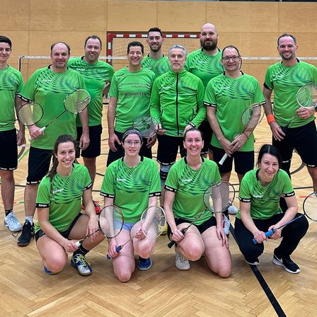 Eine Gruppe von Menschen in grünen Shirts posiert für ein Foto auf einem Badmintonplatz. Sie halten Badmintonschläger und scheinen zu lächeln. Der Platz hat Holzboden und ein Netz.
