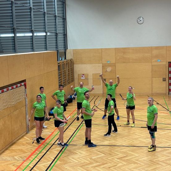 Eine Gruppe von Menschen in grünen Uniformen und Turnschuhen posiert mit Badmintonschlägern in einer Turnhalle. Einige heben ihre Hände. An der Wand hängt eine Uhr.
