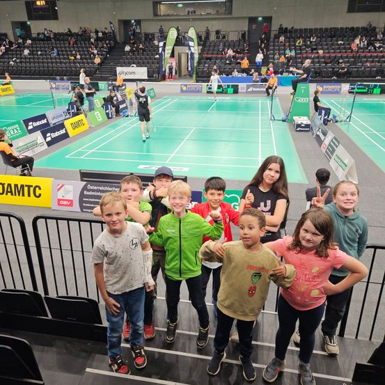 Eine Gruppe von Kindern steht im Stadion und schaut ein Badmintonspiel, mit einem leeren Spielfeld und Publikum im Hintergrund.