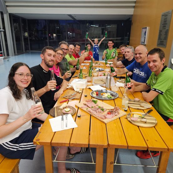 Eine Gruppe von Menschen sitzt an einem langen Tisch, lächelt und posiert für ein Foto. Sie halten Flaschen in den Händen und haben Teller mit Essen vor sich. Ein Junge steht im Hintergrund.
