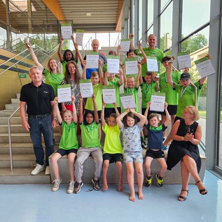 Eine Gruppe von Kindern, Erwachsenen und Trainern steht mit Urkunden auf einer Treppe und lächelt für ein Foto. Sie tragen grüne Shirts, Shorts und Turnschuhe. Hinter ihnen führt eine Treppe zu einer Glaswand, die Bäume zeigt.