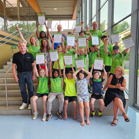 Eine Gruppe von Kindern, Erwachsenen und Trainern steht mit Urkunden auf einer Treppe und lächelt für ein Foto. Sie tragen grüne Shirts, Shorts und Turnschuhe. Hinter ihnen führt eine Treppe zu einer Glaswand, die Bäume zeigt.