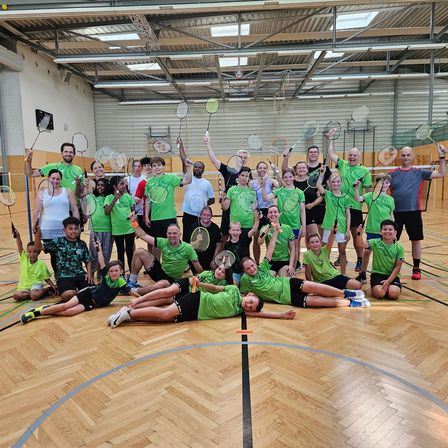 Eine Gruppe von Menschen in grünen T-Shirts und Shorts posiert für ein Foto auf einem Basketballplatz und hält Badmintonschläger in der Hand. Der Platz hat Holzböden und blaue Markierungen auf dem Boden. Die Decke ist mit weißen Lichtern und einer Uhr verziert.