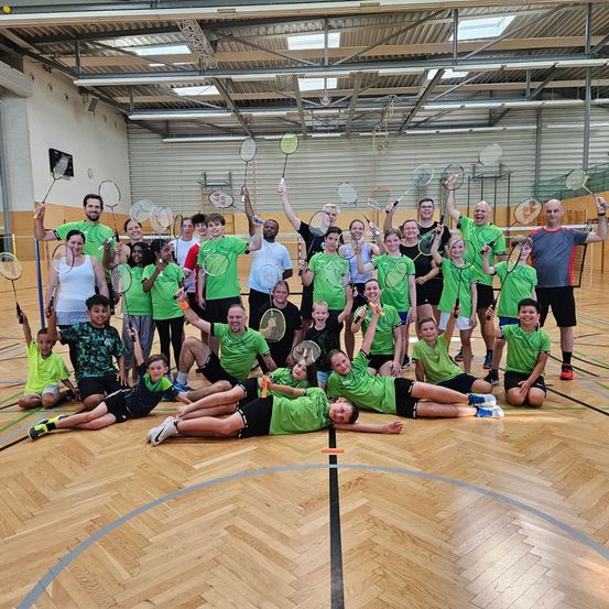 Eine Gruppe von Menschen in grünen T-Shirts und Shorts posiert für ein Foto auf einem Basketballplatz und hält Badmintonschläger in der Hand. Der Platz hat Holzböden und blaue Markierungen auf dem Boden. Die Decke ist mit weißen Lichtern und einer Uhr verziert.