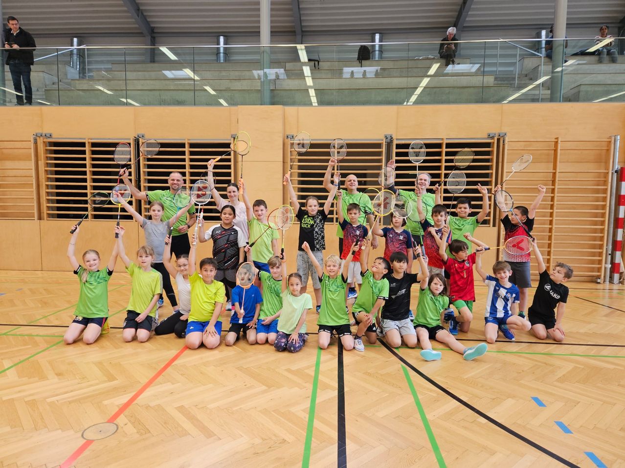 Eine Gruppe von Kindern und Erwachsenen posiert für ein Foto in einer Turnhalle mit Holzböden und hält Badmintonschläger. Die Turnhalle hat Holzwände und einen Glaszaun.