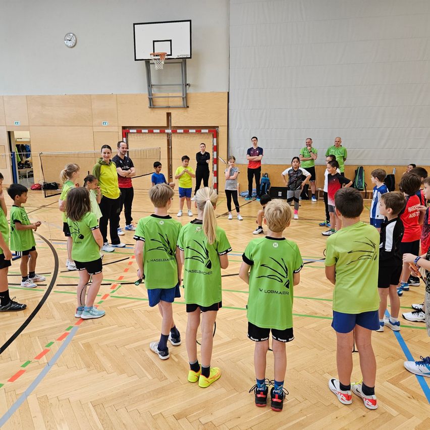 Eine Gruppe von Kindern in grünen Uniformen trainiert Basketball auf einem Hallenplatz. Mehrere Erwachsene stehen um sie herum und beobachten. Eine Uhr ist an der Wand montiert.