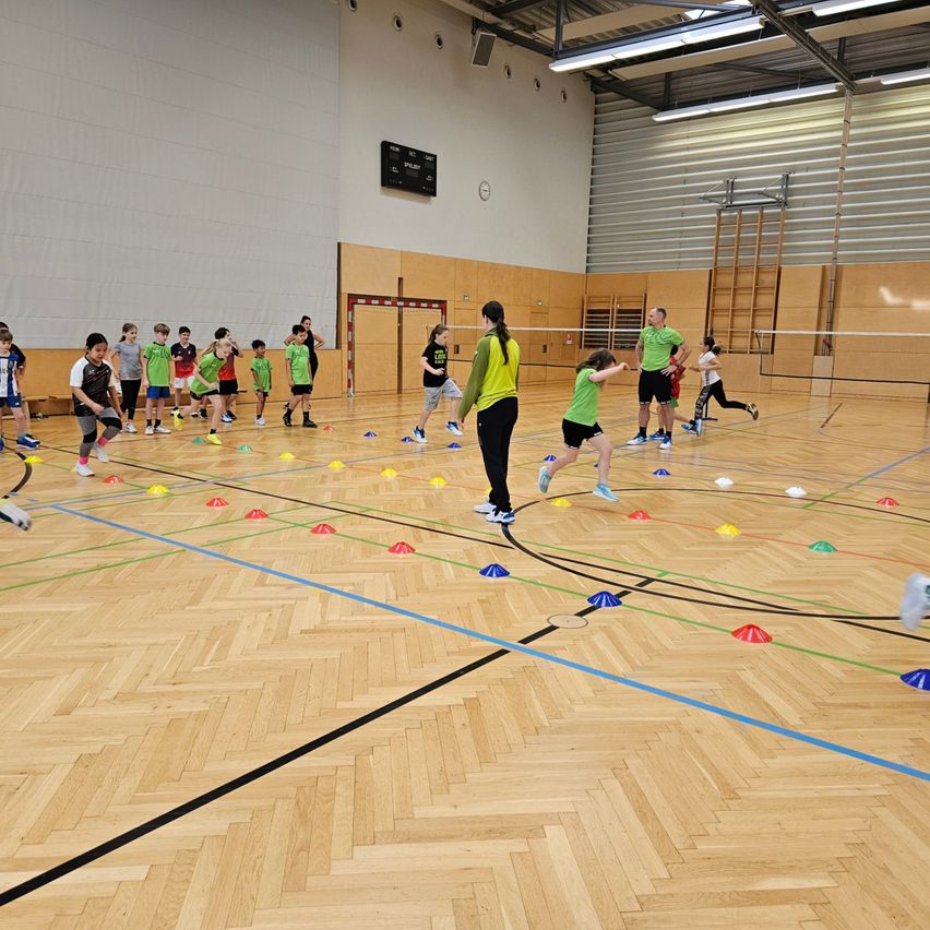 Mehrere Kinder in grünen Shirts sind in einer Turnhalle mit Holzboden an einer körperlichen Aktivität beteiligt. Erwachsene beaufsichtigen die Aktivität. Bunte Kegel sind auf dem Boden verstreut.
