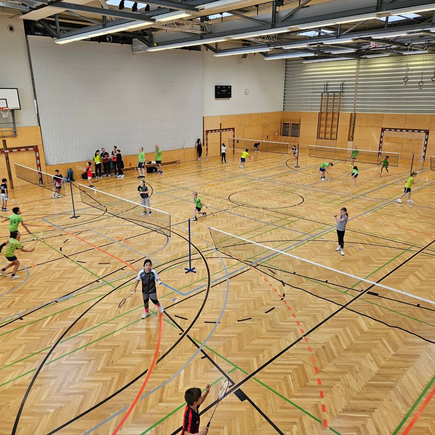 Eine Turnhalle hat mehrere Badminton-Plätze mit Spielern in grünen Trikots. Ein Spieler springt, um den Federball zu schlagen. Andere stehen und schauen zu. Die Halle hat Holzböden und Wände.