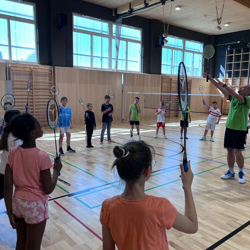 Mehrere Kinder und ein Trainer sind in einer Turnhalle und halten Badmintonschläger. Der Trainer demonstriert eine Technik.