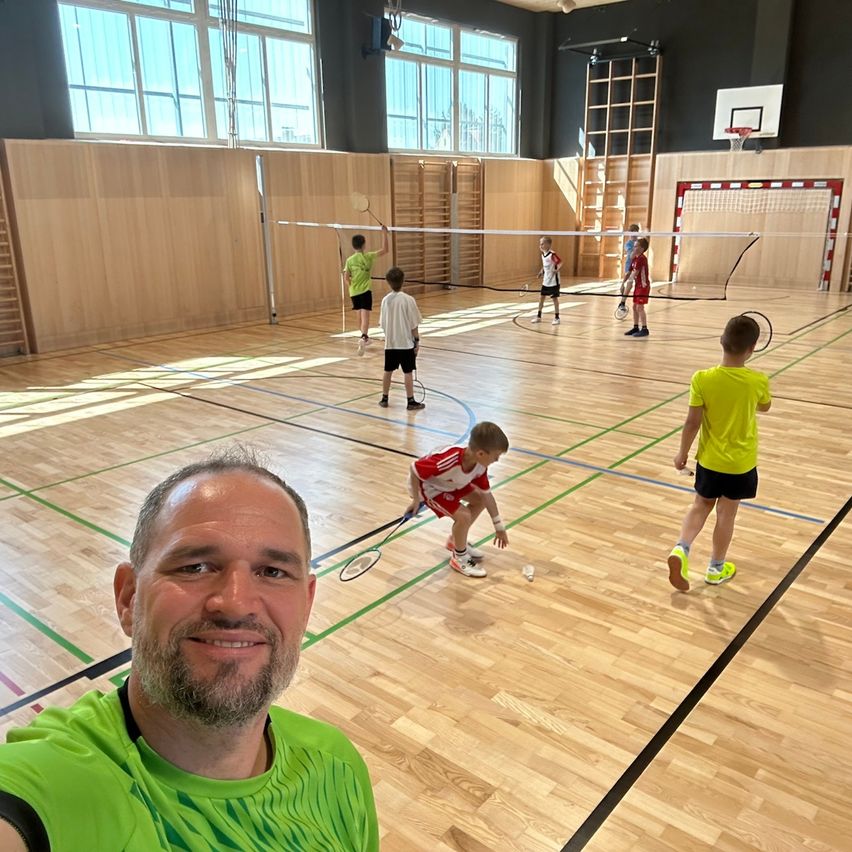Mehrere Kinder spielen Badminton in einer Turnhalle. Ein Mann lächelt im Vordergrund.
