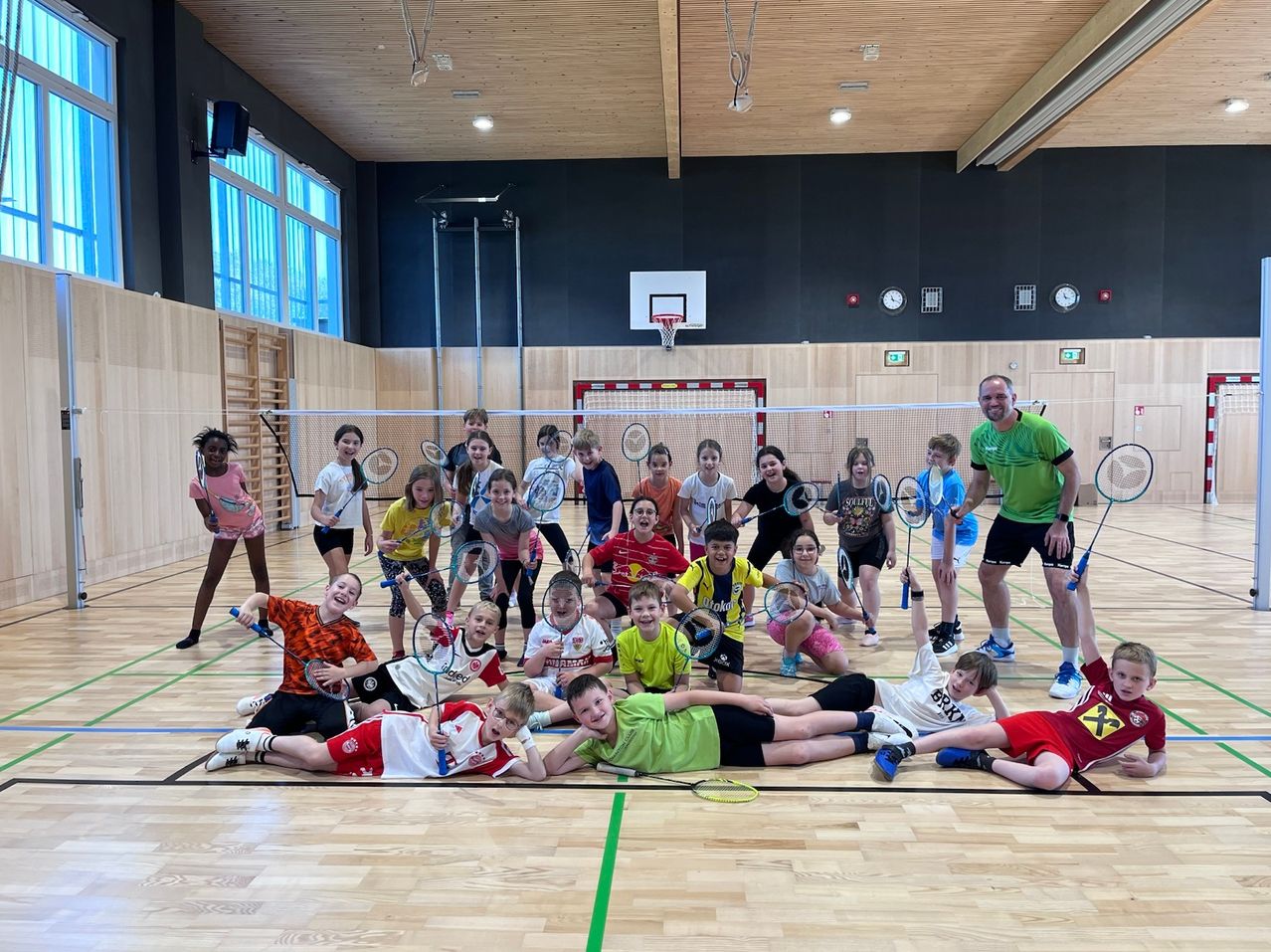 Eine Gruppe von Kindern mit Badmintonschlägern in einer Turnhalle, lächelnd und posierend für ein Foto mit einem männlichen Trainer.