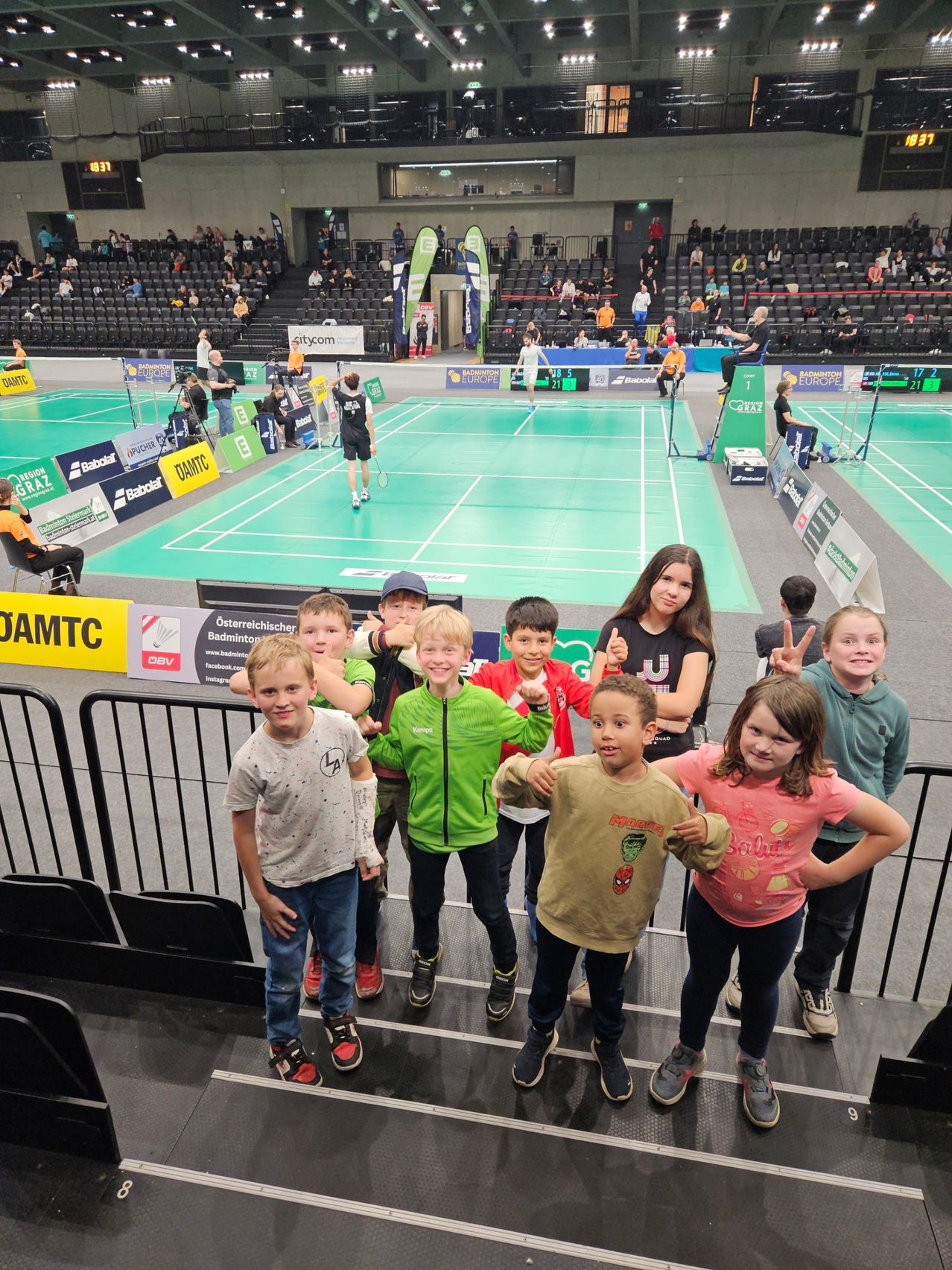 Mehrere Kinder posieren für ein Foto auf den Stufen einer Indoor-Badmintonhalle. Ein laufendes Match ist im Hintergrund zu sehen.