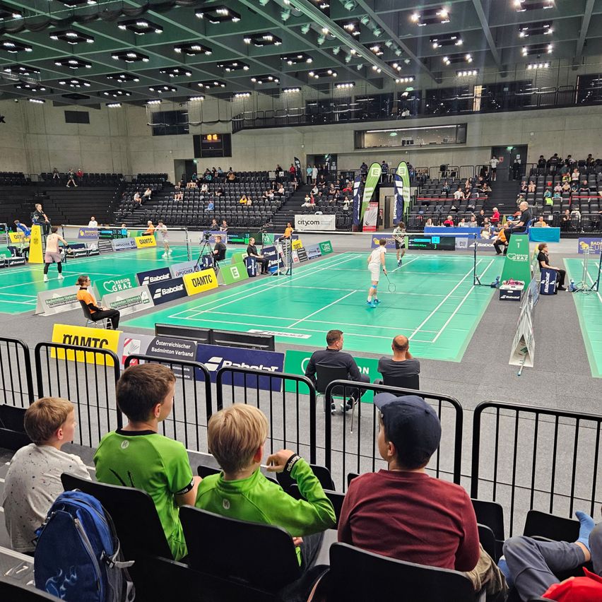 Ein Badmintonspiel findet in einer überdachten Halle statt. Mehrere Spieler sind im Spiel engagiert, während Zuschauer auf den Tribünen sitzen und zuschauen. Der Platz ist grün, und es gibt mehrere Werbetafeln und Banner rundherum.