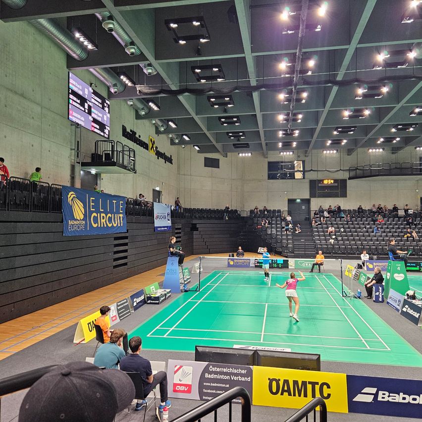 Ein Badmintonspiel findet in einer überdachten Arena statt. Spieler sind auf dem Platz, und Zuschauer beobachten von den Tribünen aus. Der Platz ist mit grünen Linien markiert, und die Spieler tragen Sportbekleidung. Banner und Werbung schmücken die Wände.