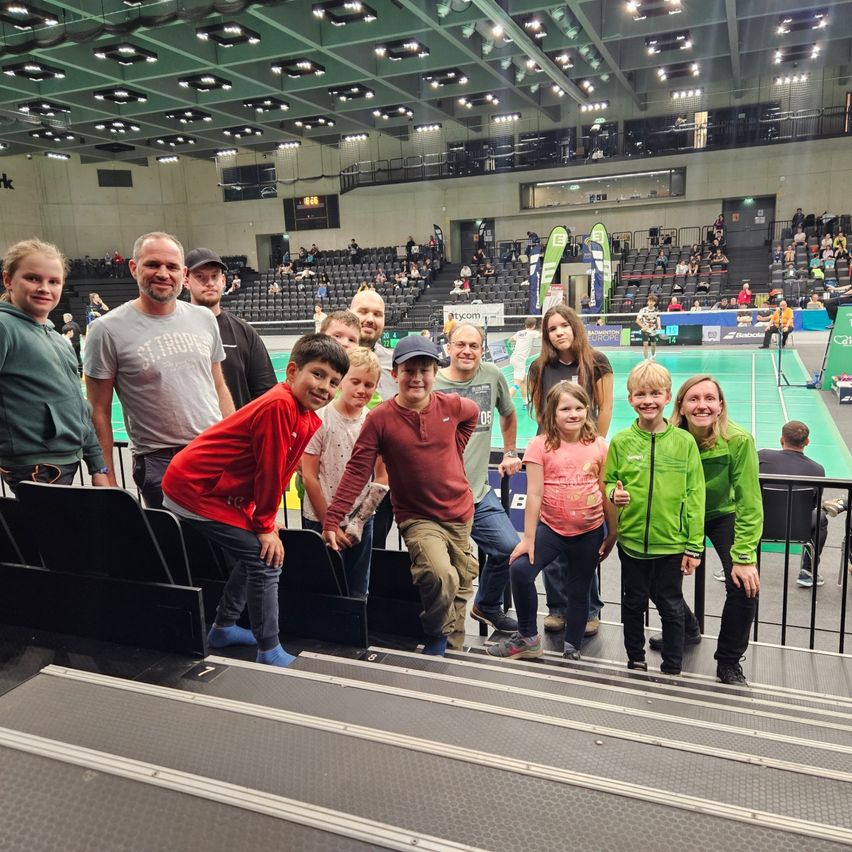 Eine Gruppe von Menschen steht auf den Stufen einer Indoor-Sportarena, lächelt und posiert für ein Foto. Es gibt Kinder und Erwachsene, einige halten Pokale. Im Hintergrund ist eine leere Arena mit einem Badmintonplatz zu sehen.