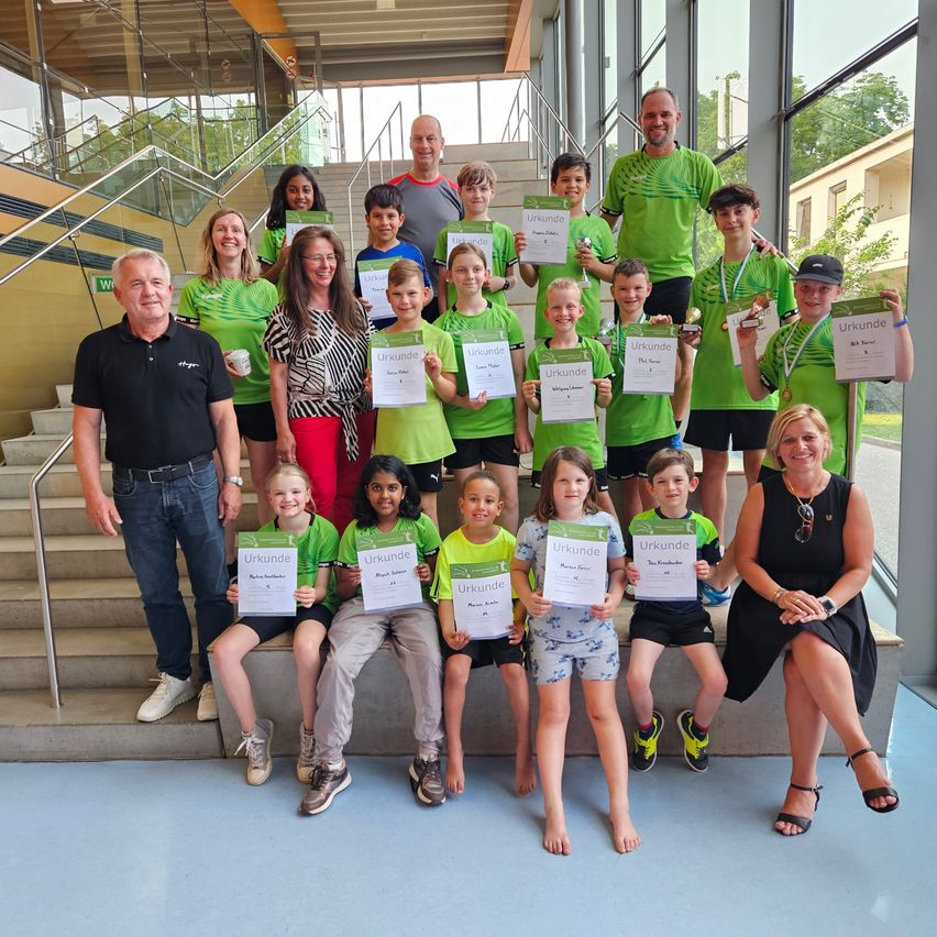 Eine Gruppe von Kindern und Erwachsenen in Grün stehen auf Stufen mit Urkunden und lächeln für ein Foto. Treppen, Glaswände und Bäume im Hintergrund.
