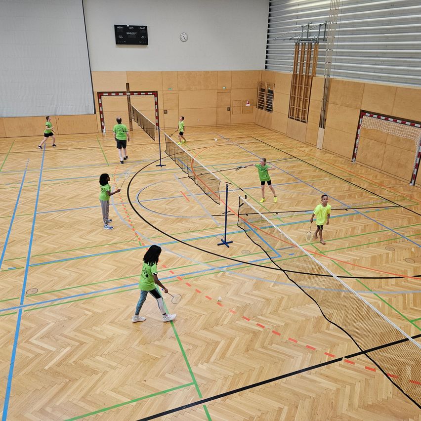 Mehrere Kinder in grünen Trikots spielen Badminton in einer Halle, markiert mit verschiedenen farbigen Linien. Der Holzboden und die Wände sind schlicht.