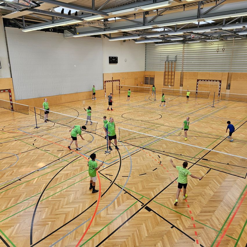 Eine Indoor-Badmintonhalle mit Holzböden und mehreren Teams von Spielern in grünen Uniformen. Der Platz ist in mehrere Bereiche mit Netzen unterteilt, und die Spieler üben und spielen.