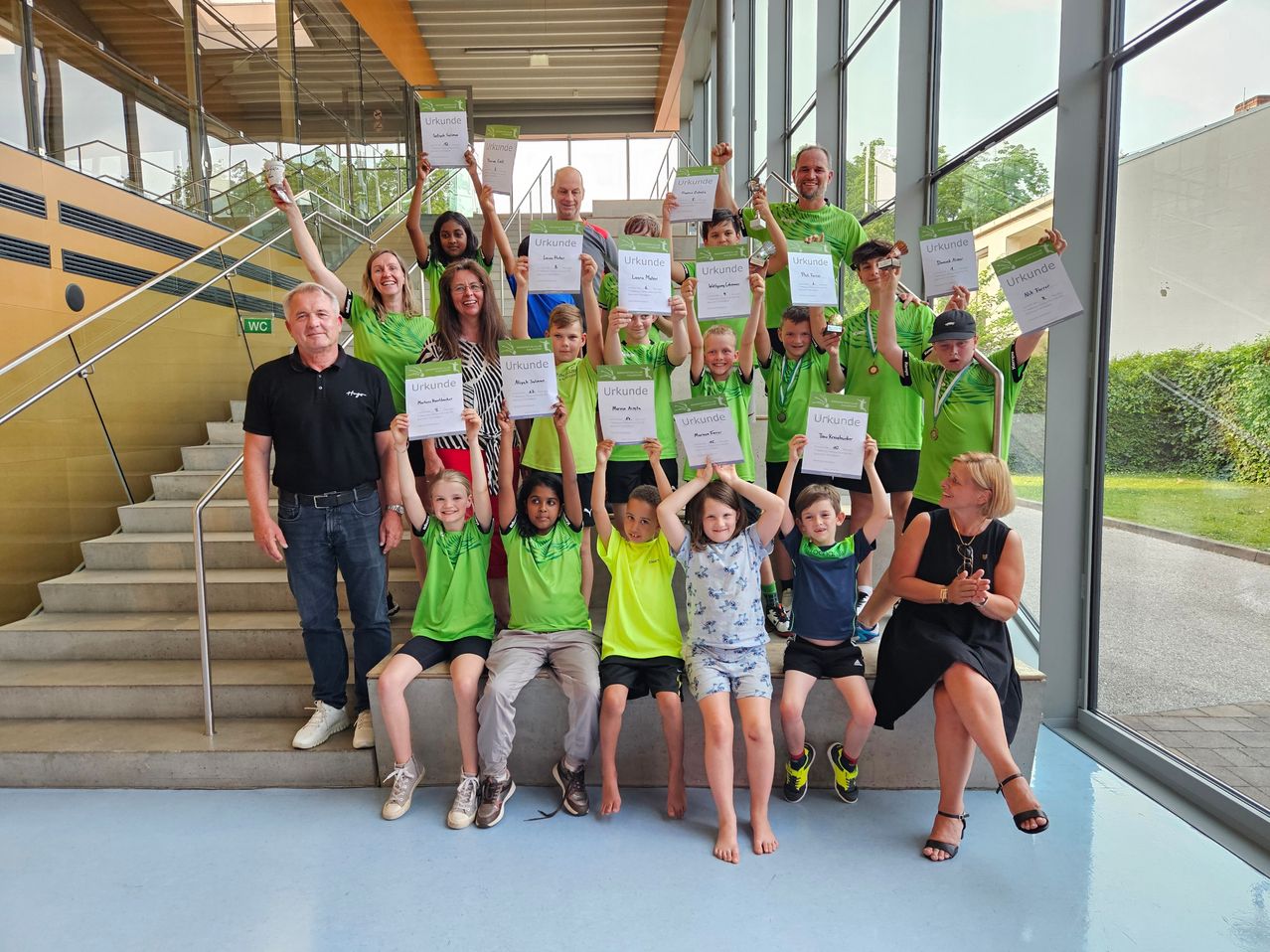 Eine Gruppe von Menschen in grünen Shirts und Shorts posiert für ein Foto mit Urkunden. Einige halten Medaillen. Sie stehen auf Treppenstufen mit einer Frau, die applaudiert. Sie befinden sich in einem Raum mit Glaswänden.
