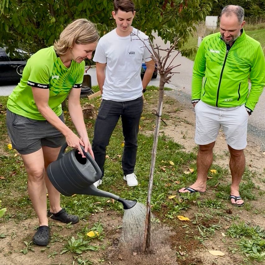 Eine Frau gießt einen Baum, während zwei Männer zusehen, einer in einem weißen T-Shirt und der andere in einer grünen Jacke.