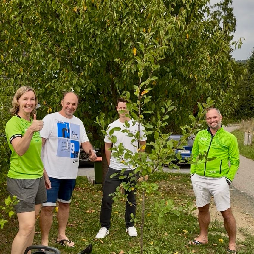 Vier Personen stehen lächelnd neben einem Baum in einem Park. Einer gibt einen Daumen hoch. Dahinter ist ein Auto geparkt. Bäume und Gras umgeben das Gebiet.