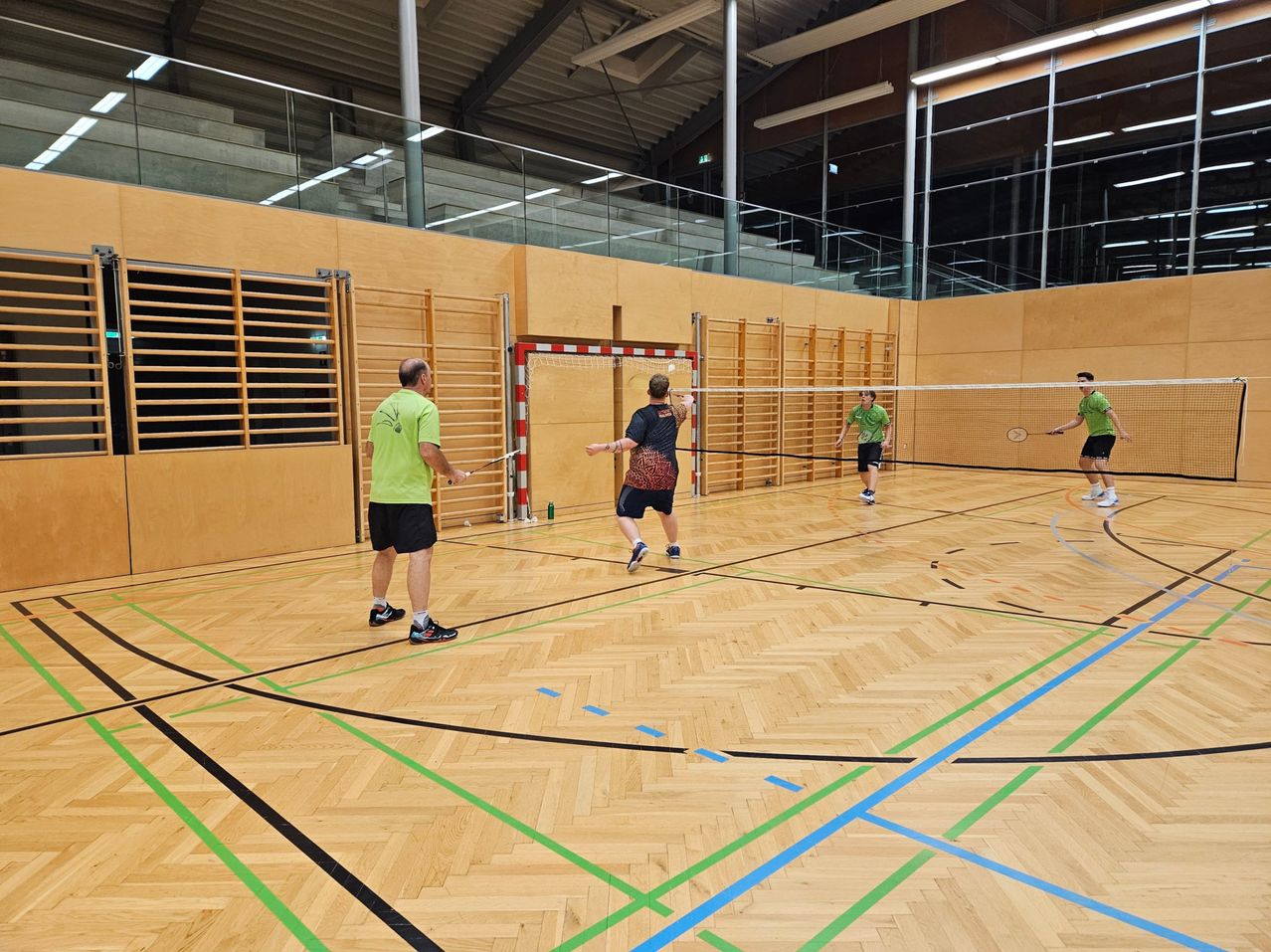 Fünf Männer spielen Hallen-Badminton auf einem mit farbigen Linien markierten Platz. Ein Mann serviert, während andere sich auf den Rückschlag vorbereiten.