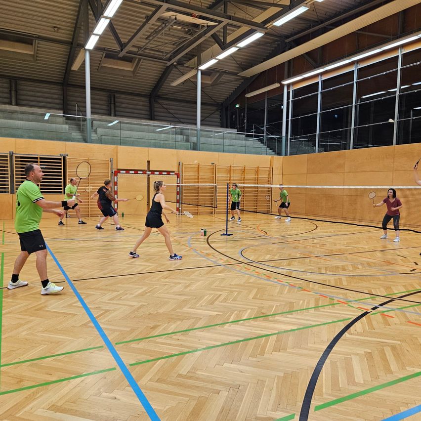 Mehrere Personen spielen Badminton in einer Indoor-Sporthalle. Sie sind in Teams aufgeteilt und verwenden Schläger und Federbälle. Der Platz hat einen Holzboden und ist mit farbigen Linien markiert.