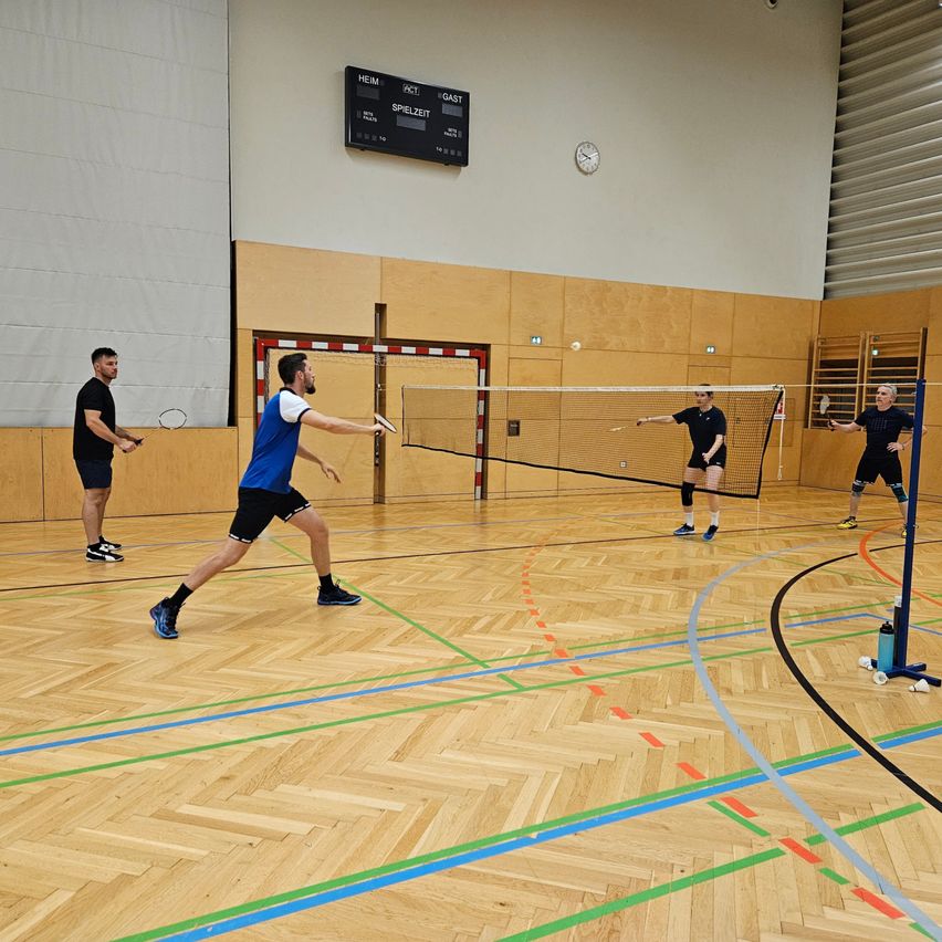 Vier Männer spielen Badminton in einer Halle. Zwei sind auf jeder Seite des Netzes, wobei ein Mann in Blau bereit ist, den Ball zu schlagen.