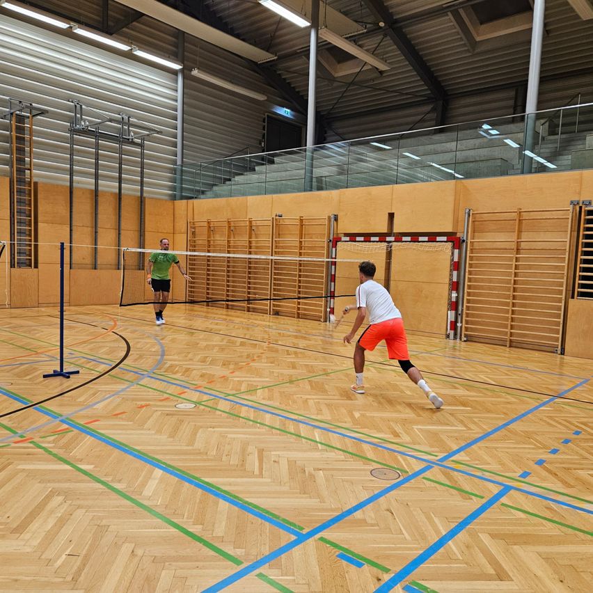 Zwei Männer spielen Badminton in einer Halle auf einem hölzernen Spielfeld mit blauen und grünen Markierungen. Ein Spieler trägt Grün, der andere Weiß. Das Spielfeld ist von hölzernen Wänden umgeben.
