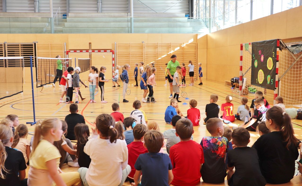 Kinder nehmen in einer Turnhalle an einer Badmintonstunde teil, einige mit Schlägern. Ein Erwachsener beobachtet. Andere sitzen auf dem Boden. Eines Tores befindet sich im Hintergrund.