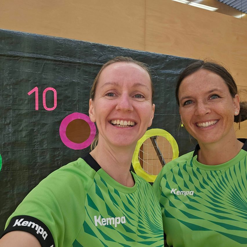 Zwei Frauen in grünen Trikots lächeln in eine Kamera in einer Sporteinrichtung. Dahinter ist eine schwarze Wand mit der Nummer 10 und bunten Ringen.