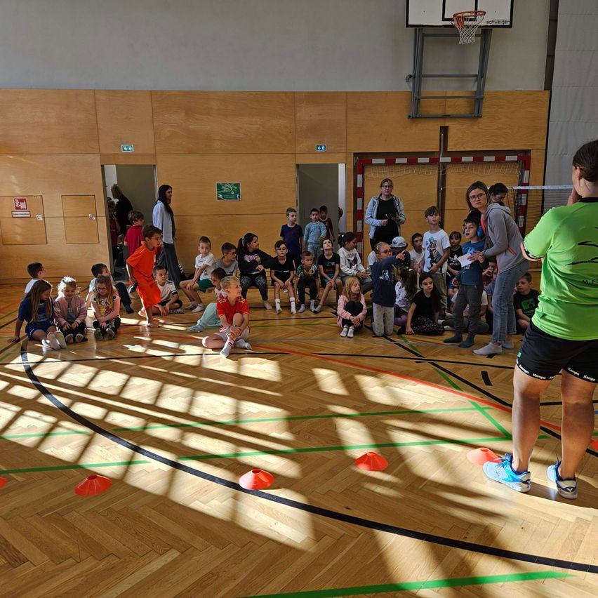 In einer Turnhalle sitzen Kinder in einem Kreis um eine Frau in Grün. Der Boden hat farbige Linien und Kegel. Ein Basketballkorb ist an der Wand montiert.