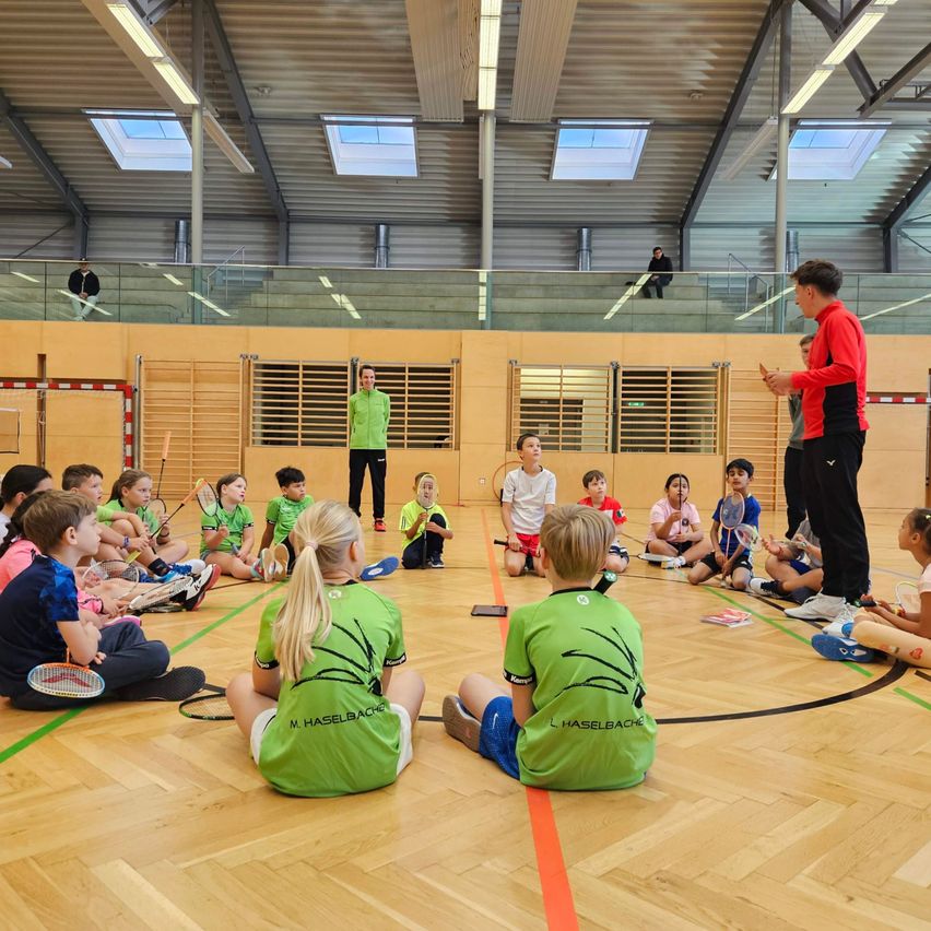 Kinder in grünen Shirts sitzen im Kreis auf einem Holzboden und hören einem Mann in einem roten Shirt zu, der vor ihnen steht. Einige Kinder halten Badmintonschläger. Im Hintergrund befindet sich eine gelbe Wand mit Fenstern und einer Balkon mit Geländer.